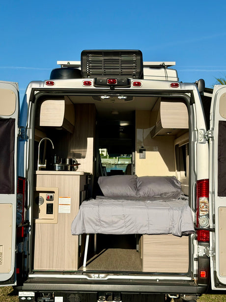 Open rear doors of this RV rental camper van show a compact interior: kitchen with sink, and a neatly made bed, ready for your trip.