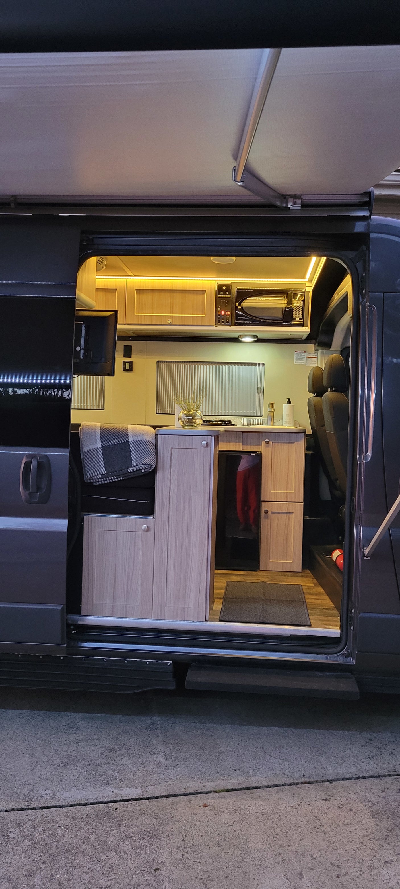 Well-lit rv rental camper van interior with kitchen, microwave, cozy seating/bed, and extended awning, ready for adventure.