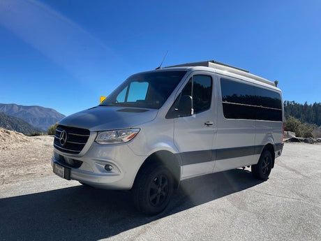 A silver Mercedes-Benz Sprinter camper van, ideal for an RV rental adventure, parked with mountains and trees under a clear sky.