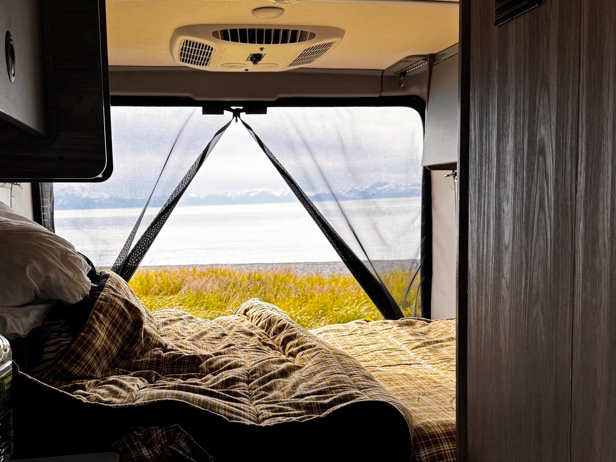  is parked on a rocky riverbed with bare trees in the background.|This gray Ram Solis rv rental camper van is parked on gravel with a wooded background — photo 5
