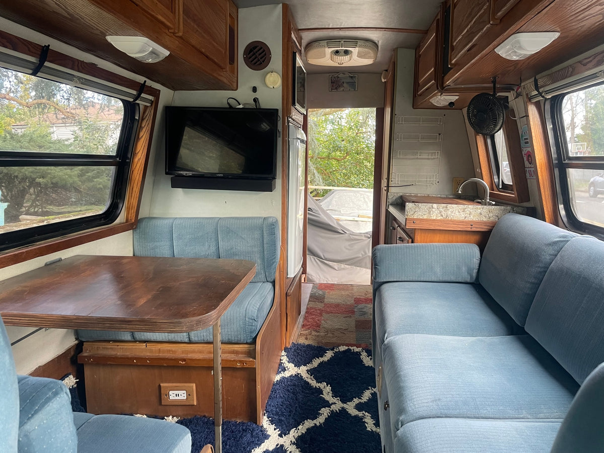 RV rental interior showcasing blue cushioned seating, a dining table, kitchen area with sink, and TV amidst wood trim. — photo 7