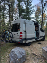 This beige rv rental camper van with bikes is parked in a natural, wooded setting, doors open for an outdoor adventure.