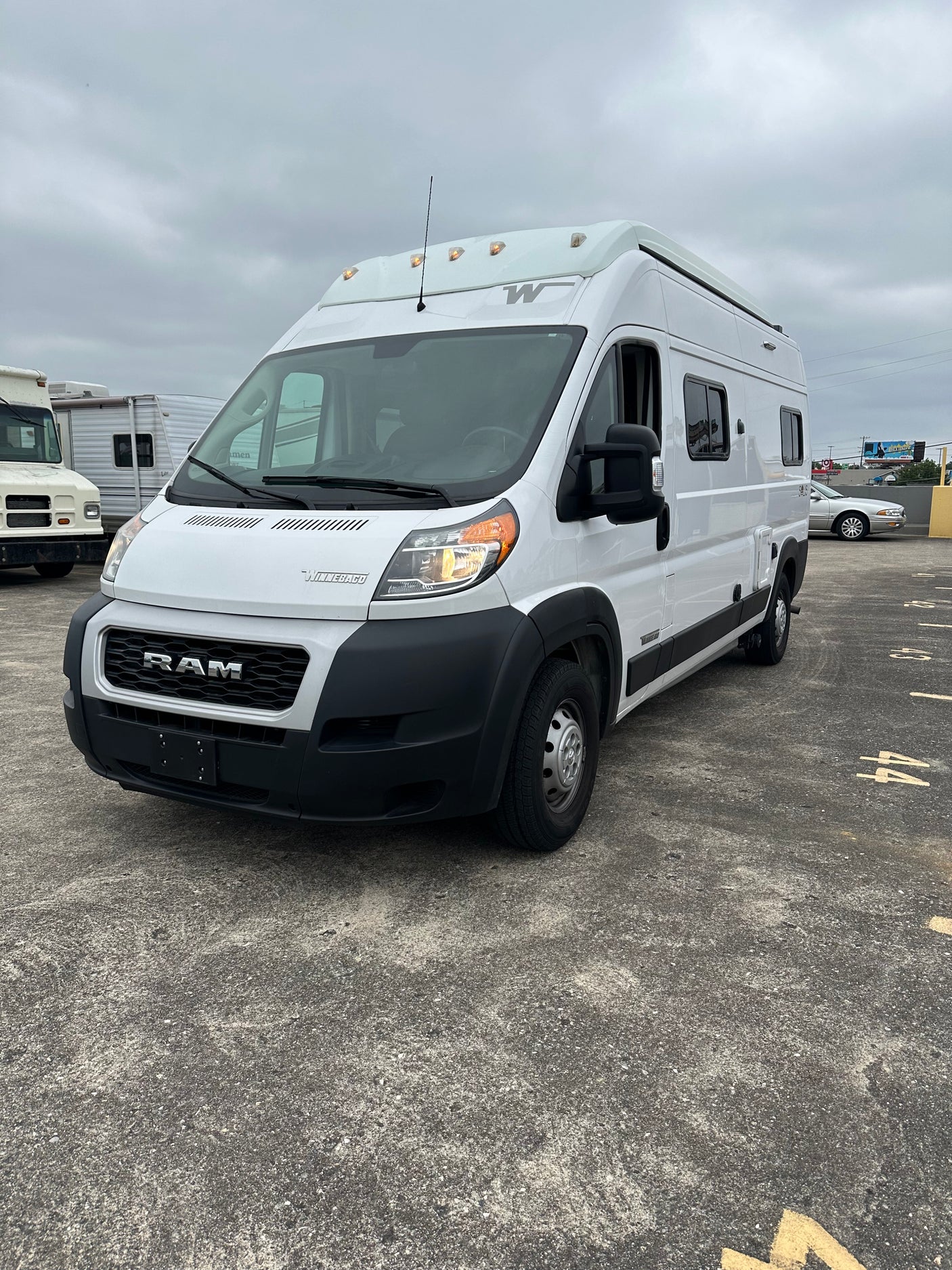 Front view of a white Winnebago Ram ProMaster RV rental van, featuring roof marker lights, parked on asphalt.