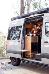 RV rental: A couple makes coffee in their well-equipped camper van kitchen, ideal for van life camping adventures with outdoor space.