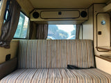 Interior view of an RV rental: striped bench seating that converts to a bed, overhead cabinets, and rear windows with curtains.