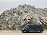 An RV rental, a dark grey Pleasure-Way camper van, is parked next to an imposing rock face, under a pale sky.