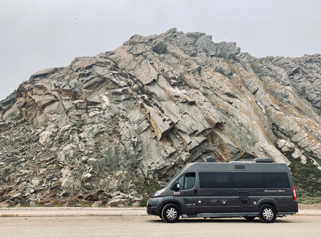 An RV rental, a dark grey Pleasure-Way camper van, is parked next to an imposing rock face, under a pale sky.