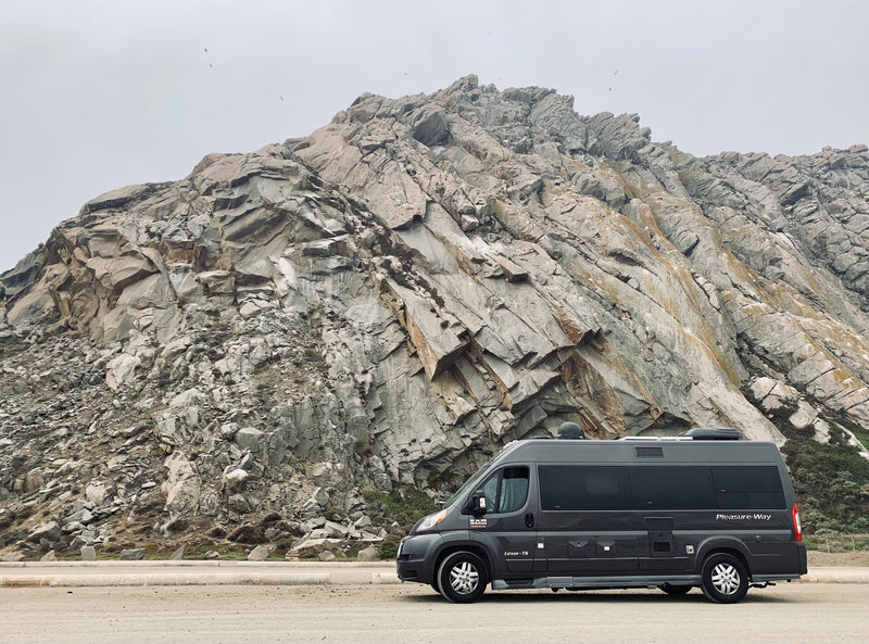 An RV rental, a dark grey Pleasure-Way camper van, is parked next to an imposing rock face, under a pale sky. — photo 5