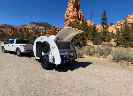 White truck tows an rv rental teardrop trailer with an open galley near stunning red rock formations under a clear sky.