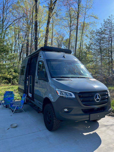 Grey Mercedes Sprinter RV rental camper van parked with open door, chairs, and fresh spring trees in a sunny outdoor setting.