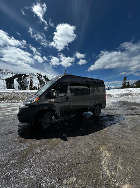 Dark grey Ram ProMaster RV rental parked on a wet mountain lot, snowy peaks & blue sky in the background. Ready for adventure!
