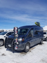An adventure-ready RV rental van, fully loaded with a snowboard and kayak, parked in a snowy mountain setting.