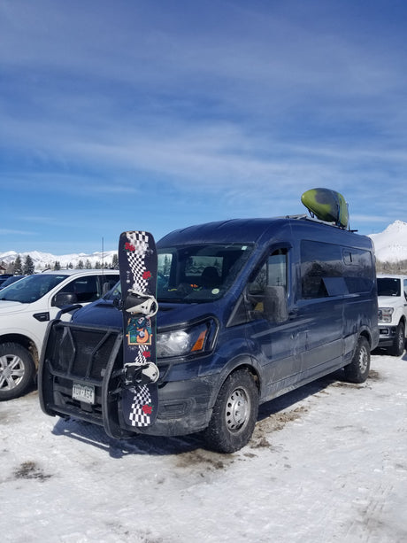 An adventure-ready RV rental van, fully loaded with a snowboard and kayak, parked in a snowy mountain setting.