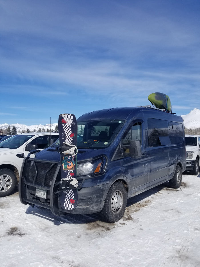 An adventure-ready RV rental van, fully loaded with a snowboard and kayak, parked in a snowy mountain setting.