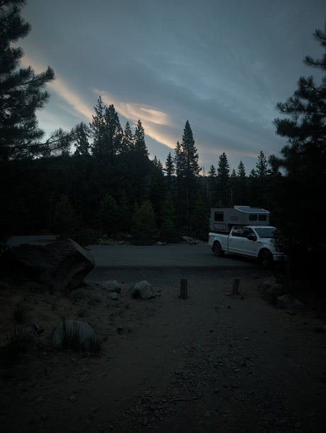 White truck with camper at a dark forest campsite at dusk. Your perfect RV rental for exploring the wilderness.