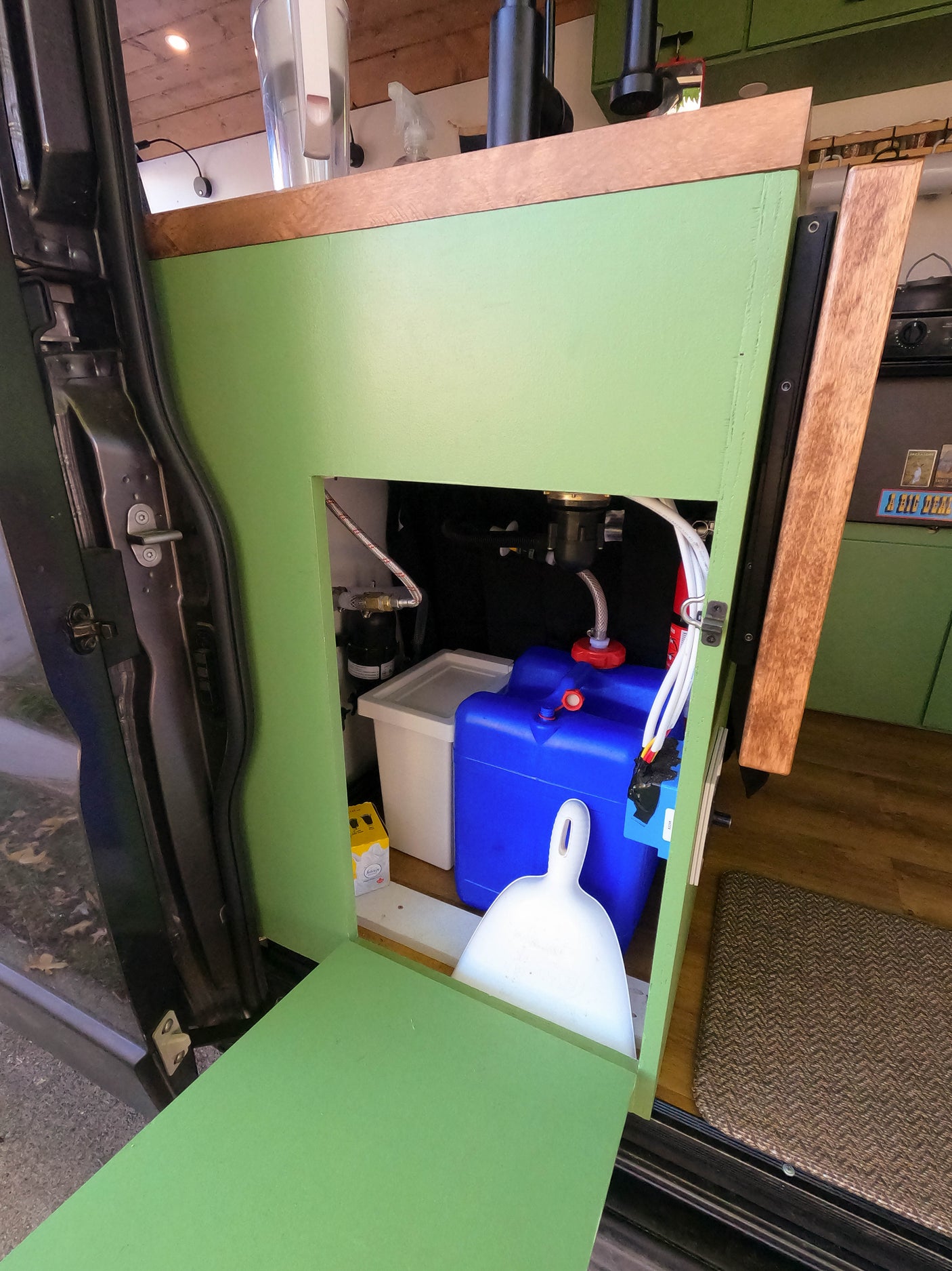RV rental interior: Green cabinet reveals blue & white water tanks & plumbing. Compact camper van living.