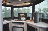 Inside an RV rental: a modern kitchen with panoramic windows, sink, stove, coffee maker, and appliances.