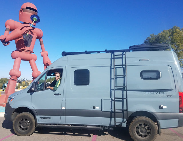 Smiling man in an RV rental (Winnebago Revel 4x4 camper van) next to a giant red robot holding a flower.