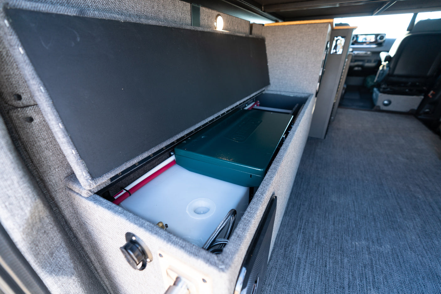 RV rental interior with open storage showing a water tank, camping stove, and plumbing, ideal for road trips.