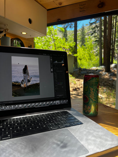 Enjoying remote photo editing in an RV rental, with a scenic forest view and an energy drink nearby.