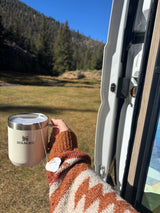 Cozy moment in an **rv rental**: Hand holding a Stanley mug with tea, admiring the forest and stream. Nature's peace.