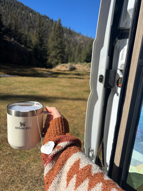 Cozy moment in an **rv rental**: Hand holding a Stanley mug with tea, admiring the forest and stream. Nature's peace.