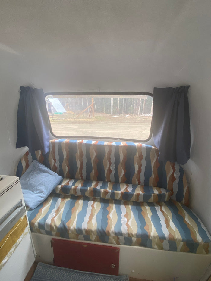 Cozy rv rental interior featuring a patterned bench/bed, window view of trees, and blue pillow. Ideal for comfortable travel.