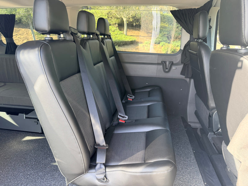 RV rental: Inside a van showing three black passenger seats, each with a seatbelt. Side windows have curtains. A bed is visible left rear. — photo 12