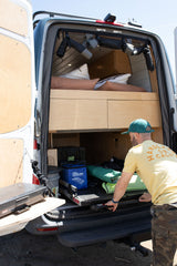 RV rental setup: Man extends a bed slide with a portable stove from a camper van, showcasing smart storage and a cozy bed.