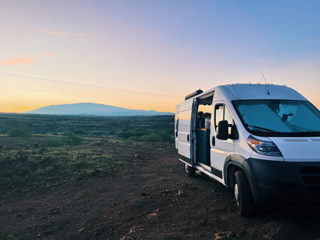 A white RV rental camper van is parked on a dirt road, overlooking distant mountains under a beautiful sunset sky.