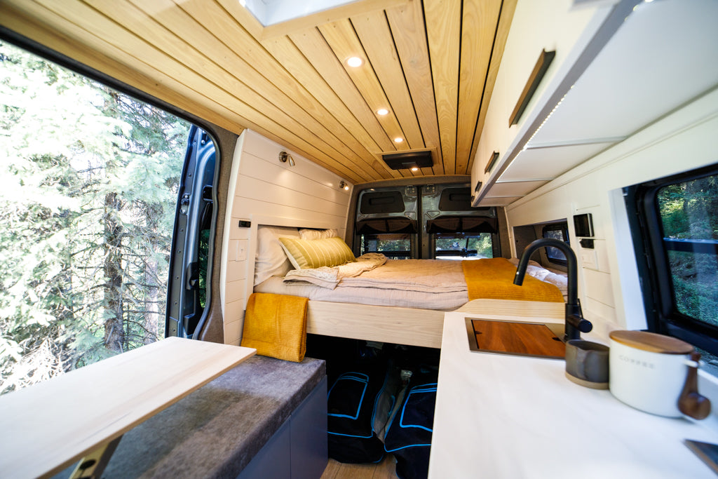 Stylish RV rental interior: wood ceiling, bed, kitchen area with sink, & stunning forest views from the open side door.