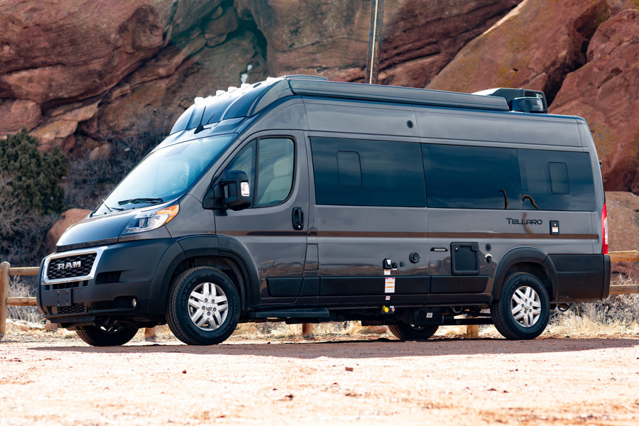 A dark gray camper van sits on a dirt road with red rock formations in the background. Ideal for an RV rental adventure.