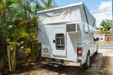 White truck with custom pop-top RV rental camper and AC unit, parked in a sunny driveway with palm trees.