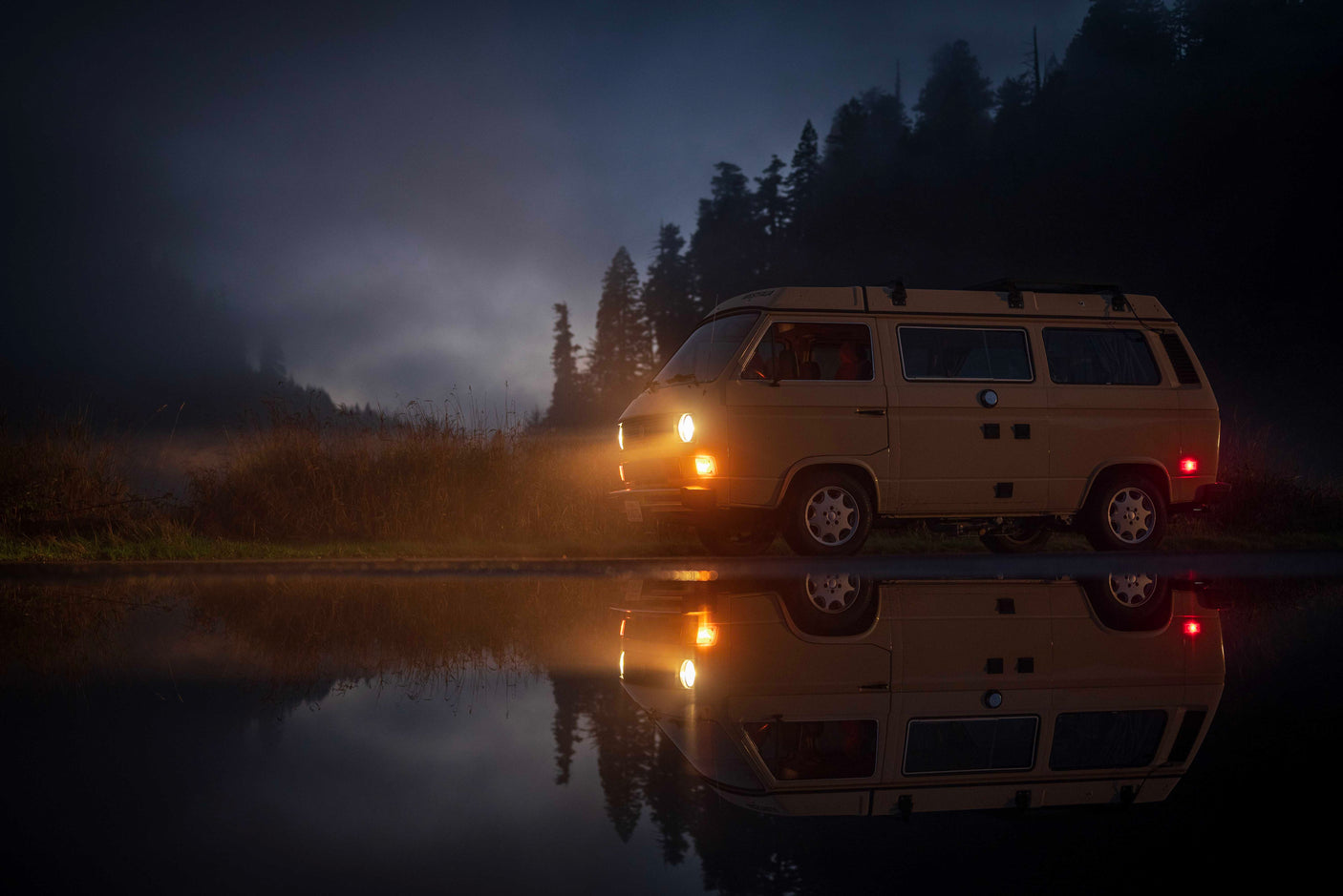 Ready for a misty night adventure? This tan VW camper van, an ideal RV rental, casts a warm glow & reflection on the wet road.