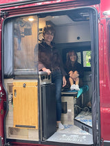 Two women inside a red RV rental camper van. One stands by the kitchen, another sits, showcasing a compact interior for travel.