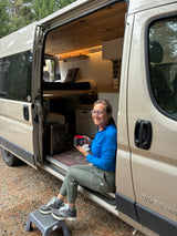 Enjoying an RV rental adventure: A woman sips coffee, smiling from her camper van's open doorway, showcasing its cozy interior.