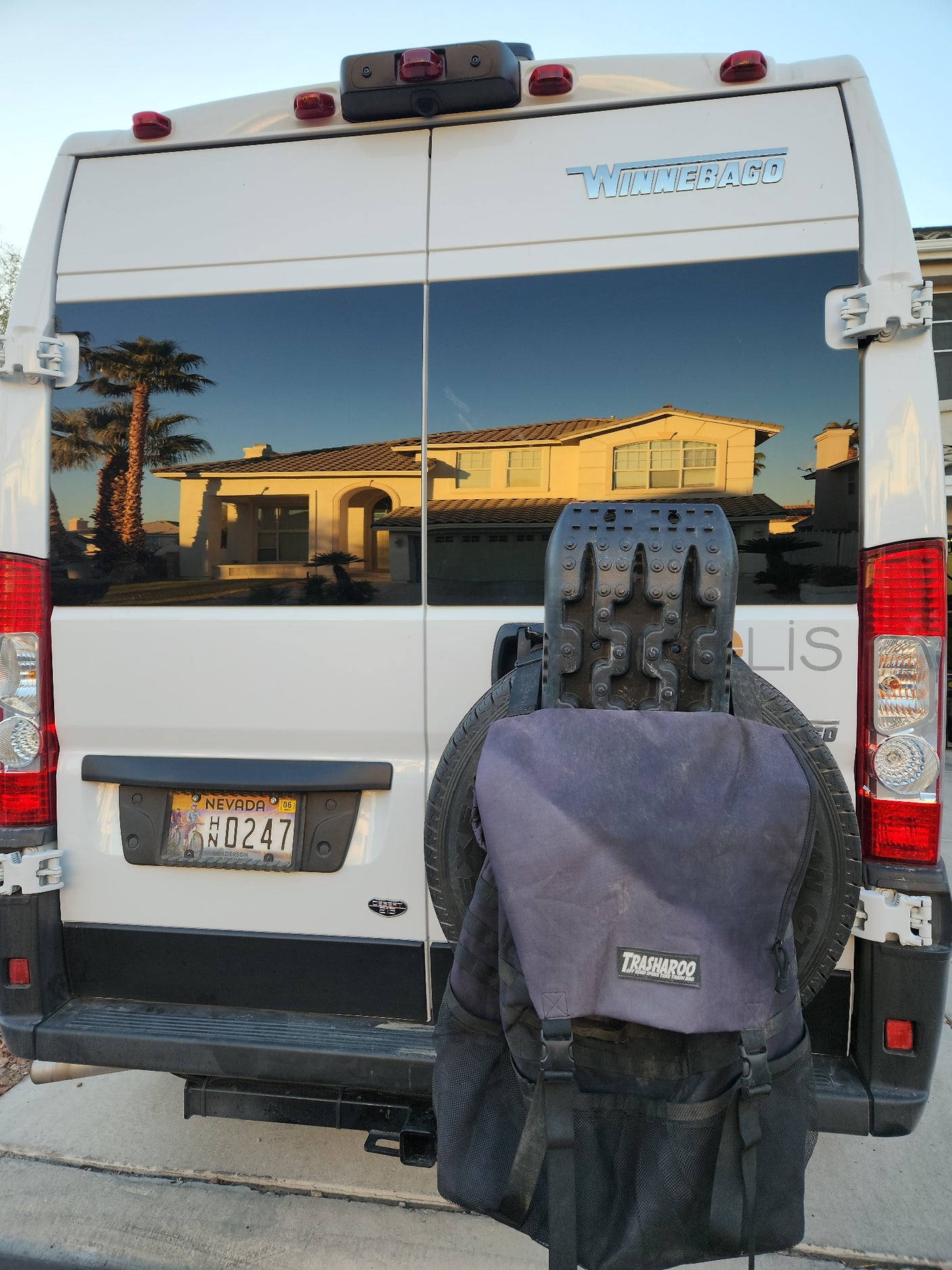 Rear of a white Winnebago rv rental van, reflecting a house & palm trees. Features a Nevada plate, spare tire, recovery board, and Trasharoo bag.