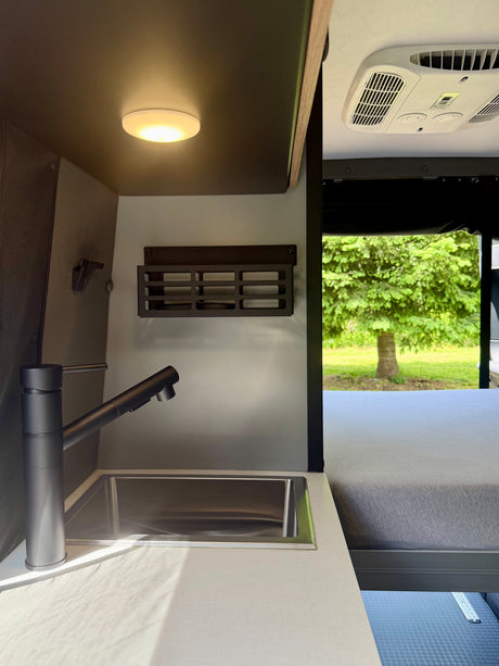 Modern RV rental interior features a sleek kitchen with a sink, faucet, overhead light, AC unit, and a cozy bed with a scenic view of trees.