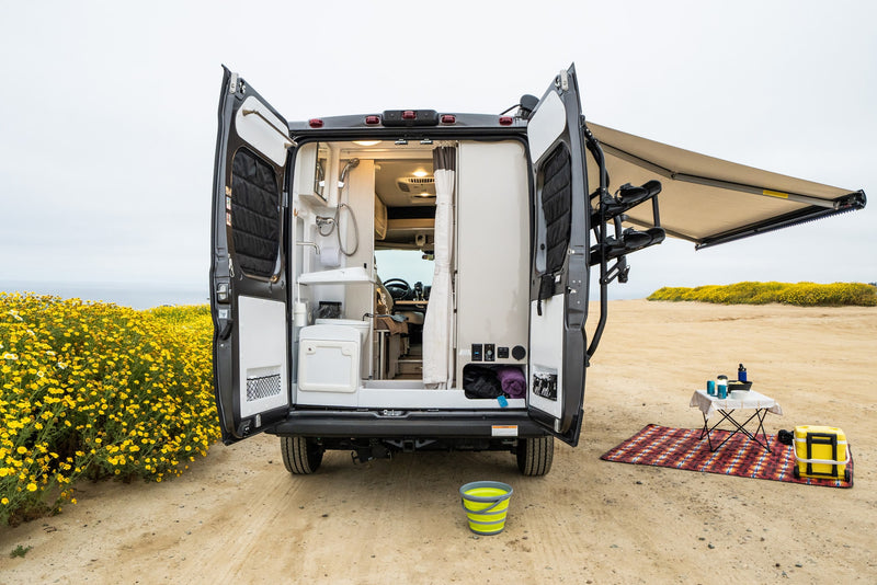 RV rental with rear doors open, showcasing interior amenities & an outdoor camping setup with table, cooler, and ocean views. — photo 14