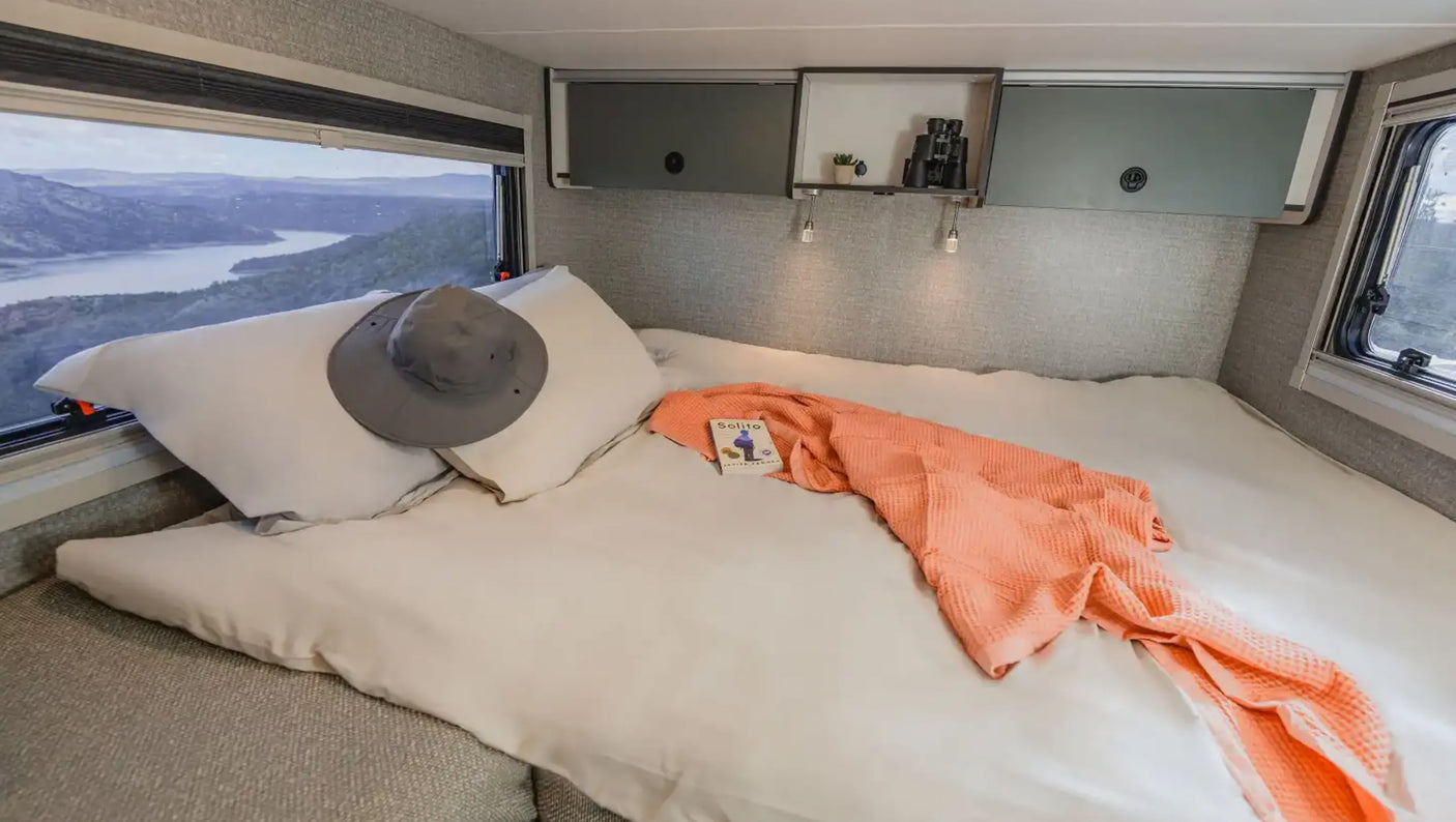 RV rental bedroom features a cozy bed with a hat & throw. A wide window offers stunning mountain and lake views.
