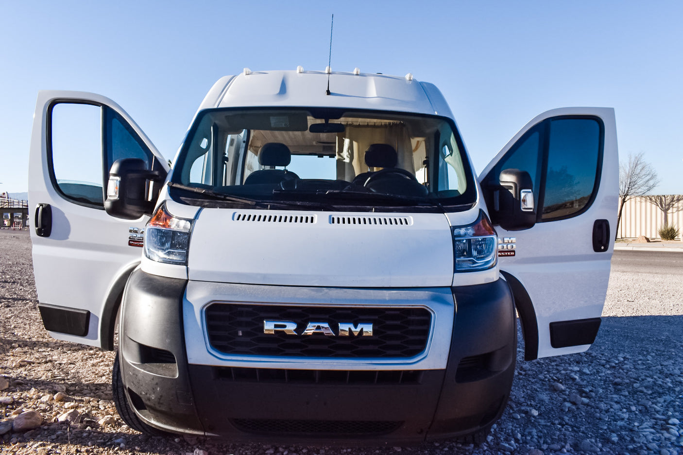 A white Ram ProMaster camper van, an ideal RV rental, with open front doors parked on gravel under a clear sky.