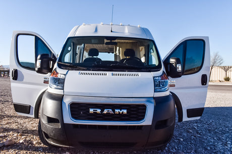 A white Ram ProMaster camper van, an ideal RV rental, with open front doors parked on gravel under a clear sky.
