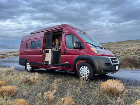 Woman inside a red Winnebago Solis RV rental camper van, parked by a lake and hills under a cloudy sky.