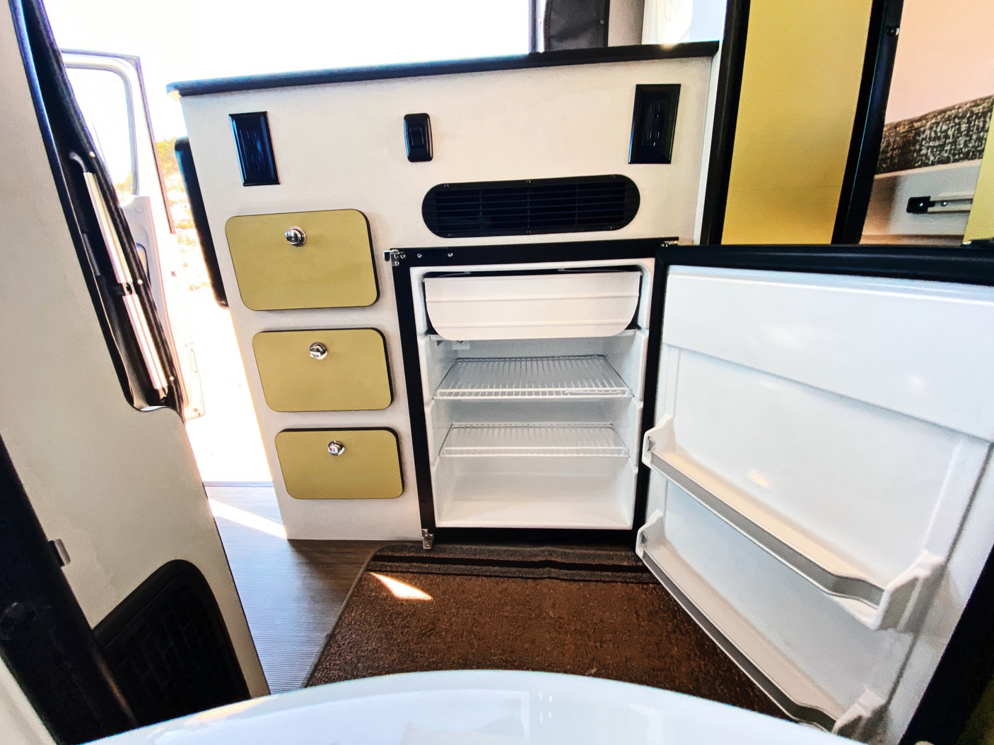 View inside an RV rental, featuring an open compact refrigerator/freezer, storage drawers, and power outlets.