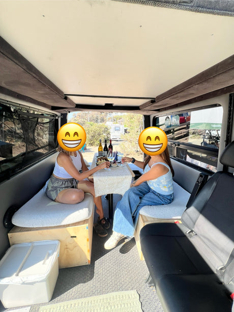 An RV rental's inviting interior: Two women toast with wine and snacks in a comfy camper van, perfect for mobile dining.