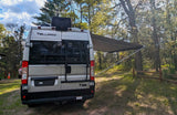 Rear view of a silver Thor Tellaro RV rental campervan with extended awning at a sunny wooded campsite, perfect for outdoor adventures.