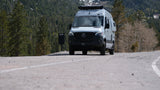 Mercedes Sprinter RV rental on a winding mountain road. Gray camper van with roof gear, surrounded by trees.