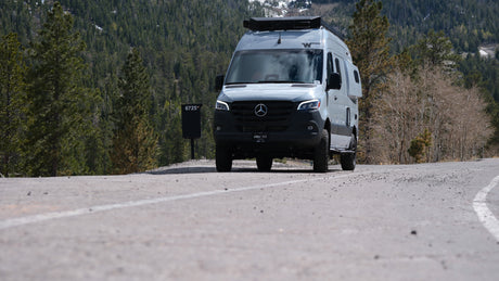Mercedes Sprinter RV rental on a winding mountain road. Gray camper van with roof gear, surrounded by trees.
