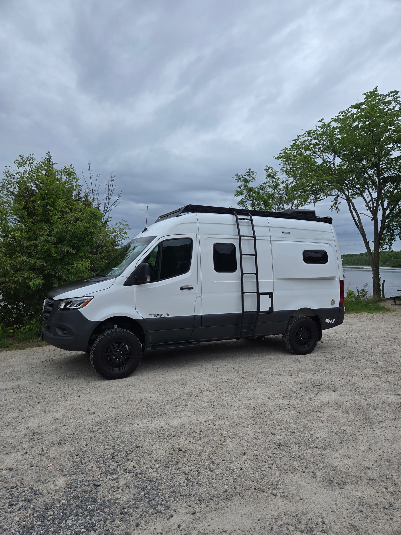 White Tiffin camper van with off-road tires and roof rack, parked on gravel near water. Ideal RV rental for adventure.