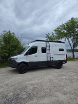 White Tiffin camper van with off-road tires and roof rack, parked on gravel near water. Ideal RV rental for adventure.